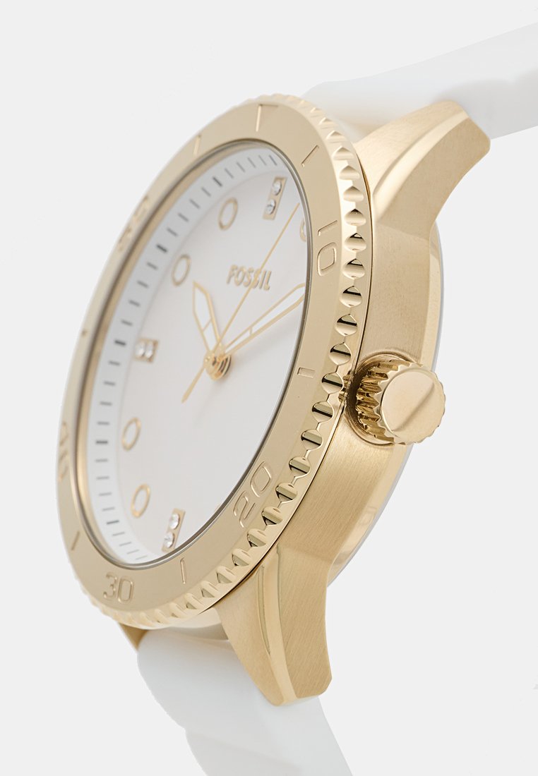 Reloj en tono dorado con una esfera blanca, marcadores de hora grabados y un bisel texturizado. Cuenta con una correa de silicona blanca y una caja de acero inoxidable.