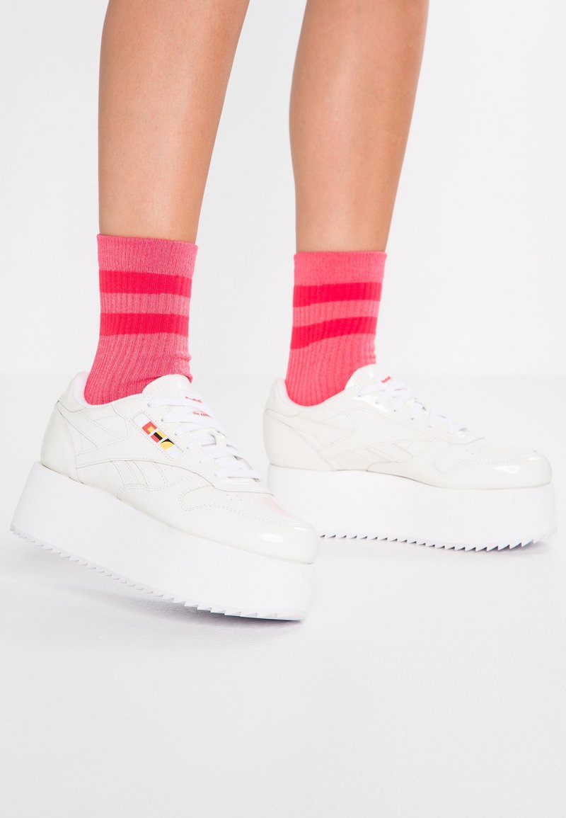 Vita plattformssneakers med texturerade gummisulor och flerfärgade accenter. Kombinerade med rosa randiga strumpor ovanför ankeln.