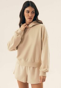 Beżowa bluzą z kapturem i długimi rękawami, połączona z dopasowanymi szortami. Gładki materiał o luźnym kroju i detalem na dolnym brzegu.