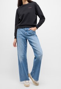 Svart sweatshirt med rosa logotyp, tillsammans med ljusblå vida jeans. Texturerad tyg, avslappnad passform, kompletterad med beige sneakers.