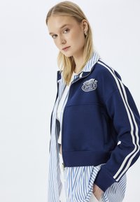 Chaqueta de cremallera azul marino con rayas blancas, logo "underground" en el pecho, combinada con una camisa a rayas azul y blanca por debajo.