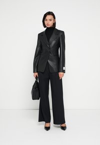 Czarna skórzana marynarka z ostrymi klapami i pojedynczym zapięciem na guzik, w połączeniu z czarnymi szerokimi spodniami i czarną torebką.