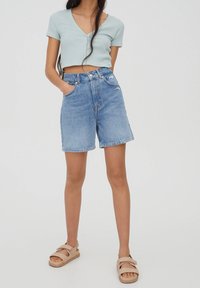 Top corto acanalado azul claro con mangas cortas y detalle de botones, combinado con shorts de mezclilla de talle alto y sandalias marrón claro.