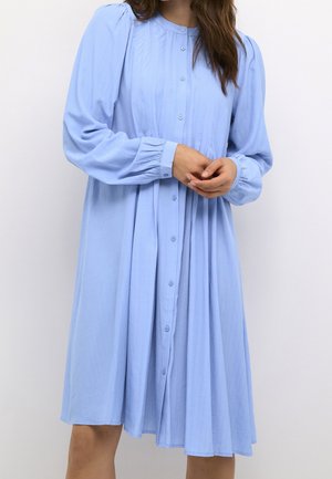 Kobieta o długich włosach, ubrana w jasnoniebieską sukienkę z przodu zapinaną na guziki, z długimi bufiastymi rękawami i plisowanymi detalami, stojąca na jednolitym białym tle.