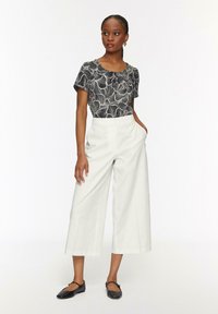 Vrouw in zwart-wit gestreept overhemd met korte mouwen, witte wijde cropped broek, zwarte platte schoenen, staande met één hand in de zak.
