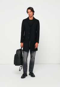 Palton lung negru cu icter, închidere cu nasturi, două buzunare frontale, purtat peste un top negru. Blugi din denim gri și pantofi negri. Rucsac negru.