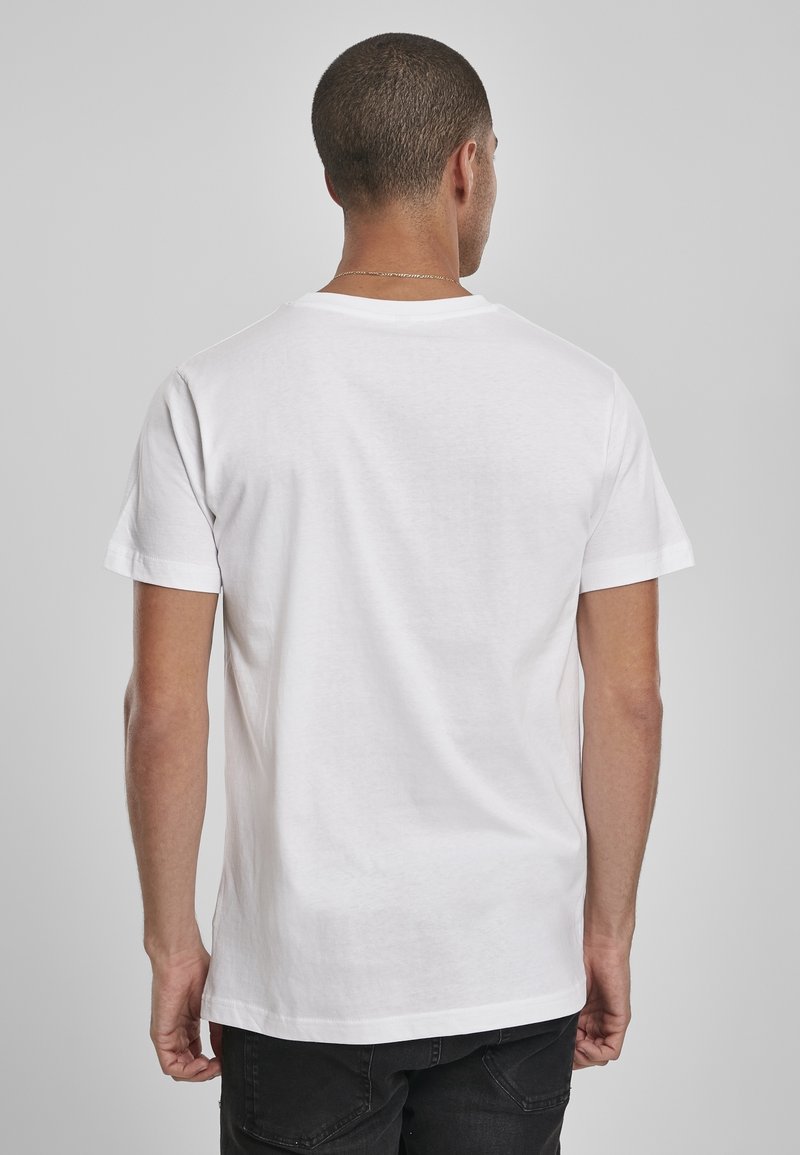 Homme portant un T-shirt blanc uni à manches courtes et un pantalon noir, vu de dos sur un fond gris uni.