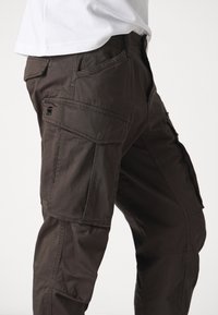 Cargo broek in donkerbruine stof met meerdere zakken, ritssluiting en een licht versleten textuur. Voorzien van logo op de zijzak.