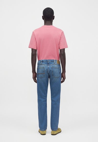 Homme debout de dos, portant une chemise rose à manches courtes, un jean bleu et des chaussures jaunes sur un fond clair uni.