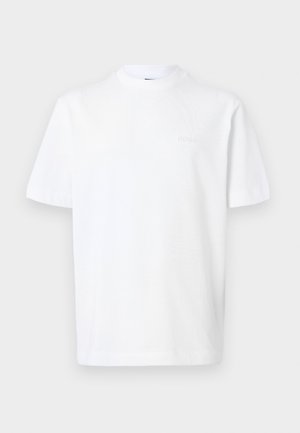 Jonge man staat tegen een witte muur, draagt een wit t-shirt, beige geplooide korte broek tot op de knie, zwarte riem, witte sokken en donkerbruine loafers.