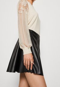 Cremefarbenes Strickoberteil mit transparenten Spitzenärmeln mit Puff-Effekt, kombiniert mit einem schwarzen Lederrock mit ausgestelltem Schnitt und glatter Textur.
