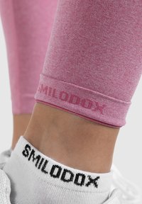 Rosa Sportleggings mit subtiler Textur, die am Saum den Markennamen "SMILODOX" in einem dunkleren Farbton aufweisen. Weiße Socken mit schwarzem Branding.