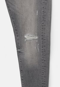Jambe de jean en denim gris avec des zones décolorées et une déchirure horizontale révélant des fils blancs.
