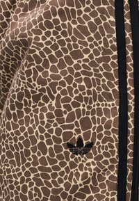Brown and beige jacket with a giraffe print pattern, featuring black accents and a small black Adidas logo. Textured fabric, zip closure.