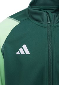 Grüne Zip-Jacke mit hellgrünen Akzenten und einer strukturierten Oberfläche. Verfügt über einen hohen Kragen und ein weißes Logo mit drei Streifen auf der linken Seite.