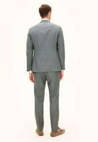 Mężczyzna w szarym garniturze i jasnobrązowych mokasynach stoi tyłem na jednolitym, jasnym tle.