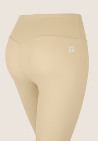 Leggings beige a vita alta realizzati in un tessuto elastico e morbido. Presentano un piccolo logo sulla parte posteriore e fianchi senza cuciture per un aspetto elegante.
