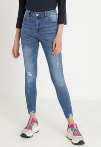Jeans ajustados en denim azul medio con detalles desgastados y dobladillos deshilachados, combinados con zapatillas deportivas multicolores que cuentan con una suela gruesa.