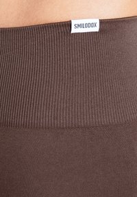 Braune, gerippte Leggings mit einem glatten Bund und einem weißen Markenlabel mit der Aufschrift "SMILODOX" obenauf. Weiche, elastische Stoffstruktur.
