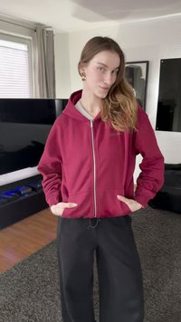 Felpa bordeaux con cerniera, cappuccio, tasche frontali e fodera interna grigia a contrasto, abbinata a pantaloni neri larghi su un pavimento testurizzato.