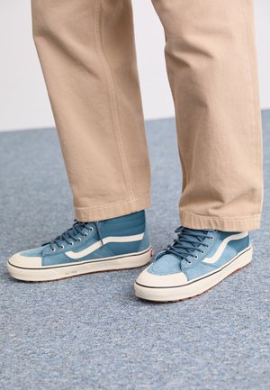 Personne portant un pantalon beige et des baskets montantes bleues avec des semelles et des lacets blancs sur un sol bleu texturé.