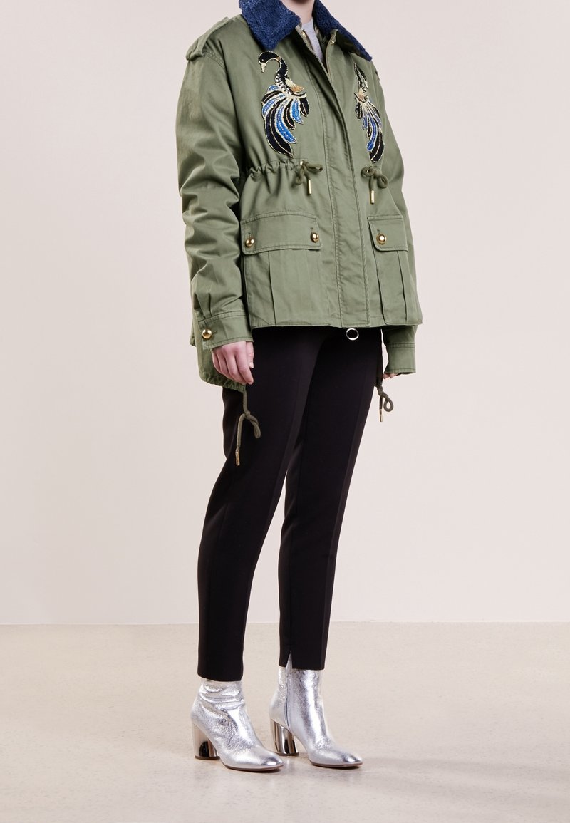 Personne portant une veste vert olive avec un col bleu et des oiseaux brodés, un pantalon noir et des bottines à talons argentés, debout.
