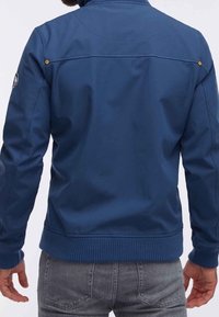 Giacca blu navy con una texture liscia, polsini e orlo a costine. Presenta dettagli gialli e una toppa con il logo sulla spalla sinistra.