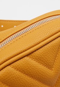 Sac en cuir jaune à surface texturée, avec une fermeture éclair argentée et des accents en forme de losanges. Vue rapprochée du matériau et du design.