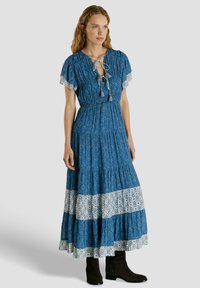 Donna che indossa un maxi abito blu con motivi a bande geometriche bianche, maniche corte, lacci con nappine al collo e stivaletti neri.