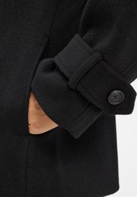 Zwarte wollenmixjas met een brede manchette met een knop, een zijzak en een zachte, gestructureerde afwerking. Het ontwerp is gestructureerd en figuurvolgend.