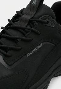 Shoe desportivo preto com uma superfície texturizada, design elegante, sistema de atacadores proeminente e um logótipo na lateral.