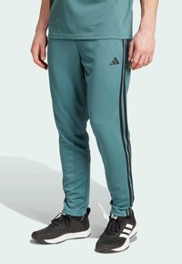 Pantalones deportivos teal hechos de un material suave, con rayas laterales negras y un pequeño logotipo; combinados con zapatillas deportivas negras con detalles blancos.