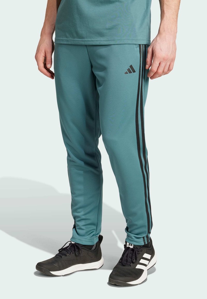 Pantalones deportivos teal hechos de un material suave, con rayas laterales negras y un pequeño logotipo; combinados con zapatillas deportivas negras con detalles blancos.