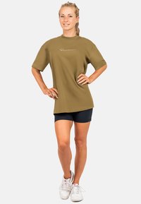 T-shirt oversize verde oliva con maniche corte e logo ricamato sul davanti, abbinato a pantaloni corti blu navy e sneakers bianche.