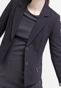 Czarny marynarka w dopasowanym kroju z pojedynczym zapięciem na guzik, klapami oraz kieszenią na suwak z boku, zestawiona z fakturowaną czarną sukienką.