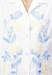 Camicia bianca con ricami floreali blu e gialli. Presenta una chiusura con bottoni e un tessuto morbido con una texture liscia.