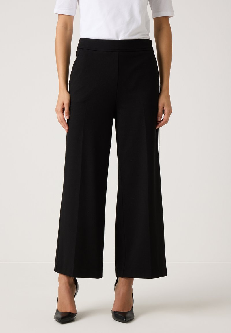 Zwarte wijde broek met een gladde, stretchy stof en een elastische tailleband, gecombineerd met zwarte hoge hakken.