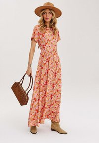 Kvetinové maxi šaty v ružovej farbe s oranžovými a červenými potlačami; krátke rukávy, zúžený pás, splývavá sukňa. Doplněné hnedou kabelkou a klobúkom.