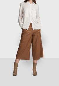 Biała koszula z długim rękawem i dwoma kieszeniami na piersi, w zestawieniu z brązowymi szerokimi skórzanymi culottes i beżowymi botkami za kostkę. Gładka tekstura i luźny krój.