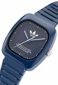 Montre en plastique bleu marine avec un boîtier carré, un bracelet texturé et un cadran présentant des aiguilles argentées et un logo blanc à trois rayures.