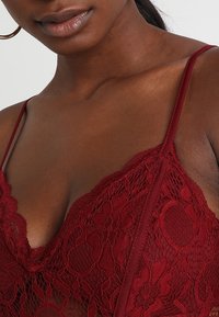 Pitsinen bralette syvän viininpunaisena, jossa on kukkakuviot, säädettävät hihnat ja V-kaula-aukko. Hento läpinäkyvä materiaali korostaa visuaalista yksityiskohtaisuutta.