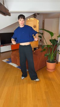 Modra kratka majica z logotipom, črne široke hlače in srebrni čevlji, postavljeno v sobo z lesenim podom in zeleno rastlino.