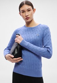 Gaiši zils džemperis ar kabeļu rakstu, ar ribas apakšu un aprocēm. Modelis tur nelielu melnu rokassomiņu. Fonā ir vienkāršs balts fons.