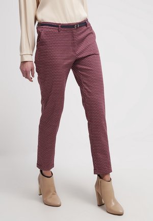 Mujer con pantalones ajustados de estampado granate, blusa beige y botines de tacón alto beige, posando contra un fondo liso.