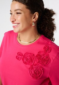 Femme souriante avec des cheveux bouclés portant un haut rose vif décoré de roses en tissu, des boucles d'oreilles en or et un collier en chaîne doré.