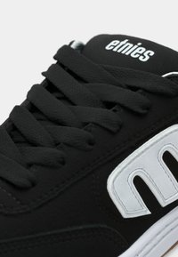 Μαύρο αθλητικό παπούτσι με μαύρες κορδόνες, λευκή σόλα και μεγάλο λευκό λογότυπο "M" στο πλάι; η επωνυμία "etnies" στην γλώσσα.