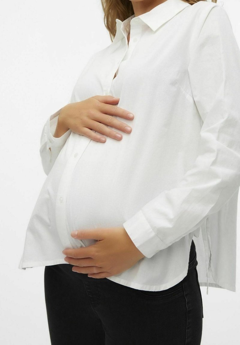 Chemise blanche à boutons, ample, à longues manches et fentes latérales, portée sur un pantalon noir, mettant en valeur un ventre de femme enceinte.