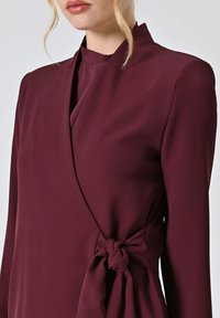 Vestito avvolgente color bordeaux in tessuto liscio, caratterizzato da maniche lunghe, colletto alto e una cintura annodata in vita per una silhouette aderente.