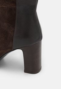 Primer plano del tacón de una bota de tobillo de cuero marrón y ante con diseño de bloque apilado sobre fondo blanco.