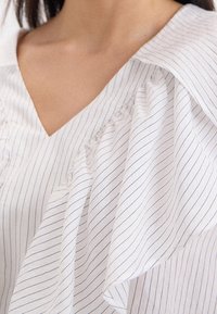 Hvid bluse med tynde lodrette striber, der har V-udskæring og flæsedetaljer langs skuldre og brystområde.